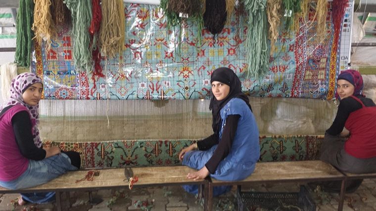 Three women sitting on a bench in front of a tapestry they are weaving.