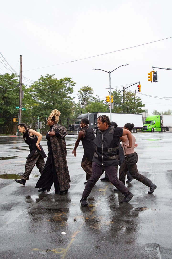 A group of performers dressed in black, dancing across a street slick with rain.
