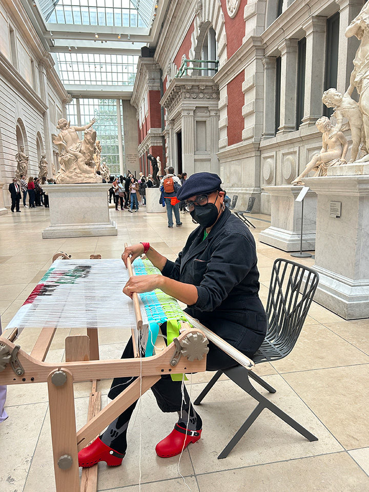 Person poses for the camera while weaving at a loom in the light-filled space of The Met's Gallery 548.