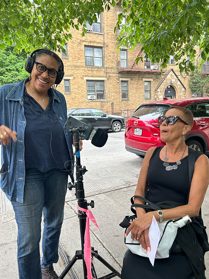 Woman in headphones stands smiling next to a microphone positioned towards a seated woman. They are outside, beneath a tree.