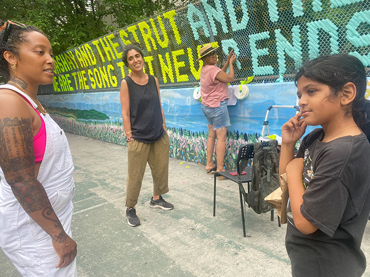 Three people speaking in the foreground, while someone in the background works at work weaving ribbon between the wires of a chain-link fence to spell out a message.