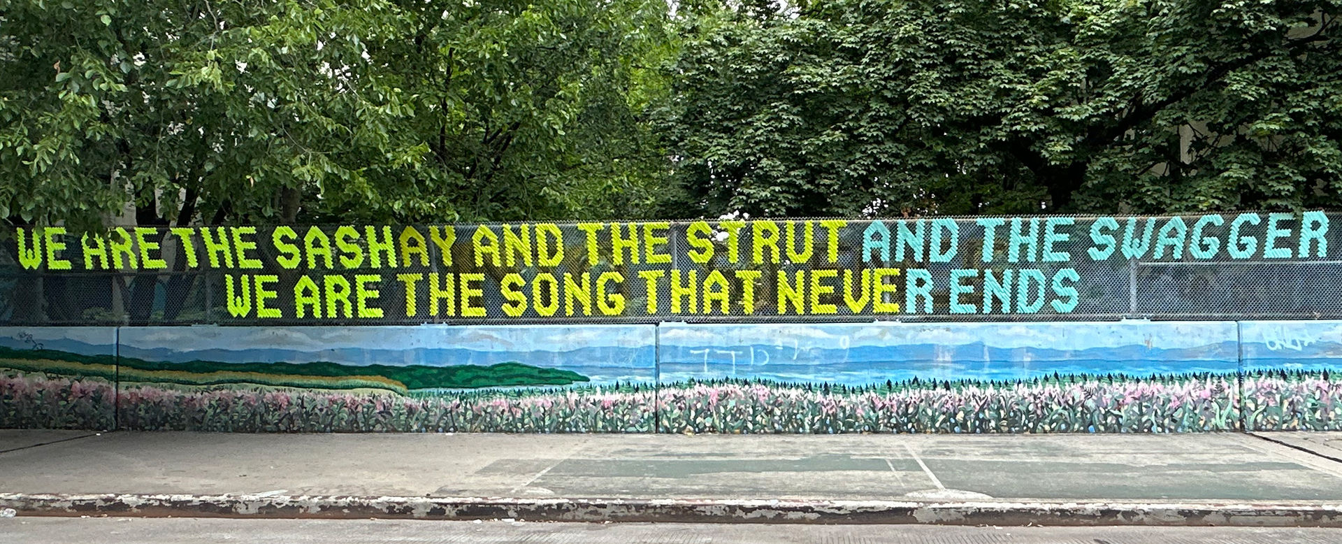 A chain-link fence is decorated with a message woven in multicolored letters: "WE ARE THE SASHAY AND THE STRUT AND THE SWAGGER / WE ARE THE SONG THAT NEVER ENDS"