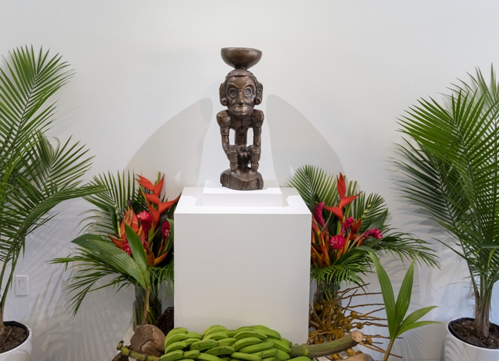 A bronze version of the Taíno Zemí replica sculpture appears on a pedestal surrounded by palm plants at the pop-up exhibition at Hope Community’s La Galeria del Barrio.