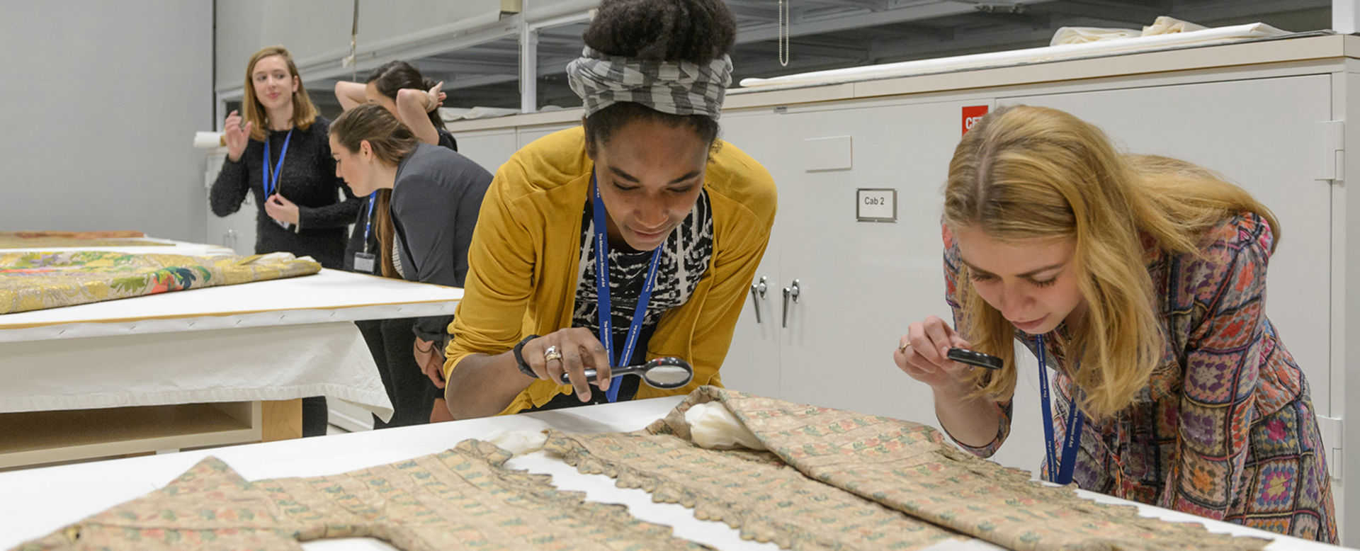 Two young women use magnifying glasses to inspect a textile.