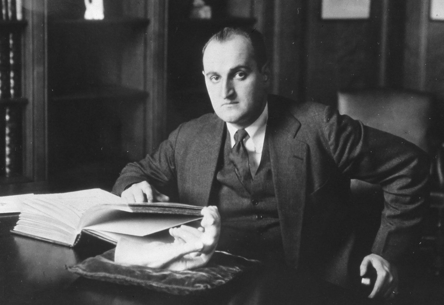 Black-and-white photograph of a man seated at a desk with a book and a sculpture of a hand. The man looks directly at the camera.