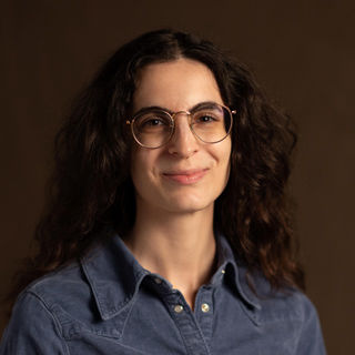 Portrait of a white woman with long, dark curly hair, smiling softly, wearing round glasses and a denim shirt.