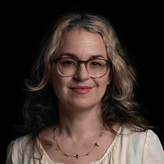 Woman with wavy brown highlighted hair and glasses, wearing a white dress, in front of a black background