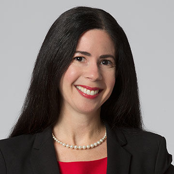 A smiling woman with long, straight dark hair, wearing a dark jacket over a red blouse with a pearl necklace