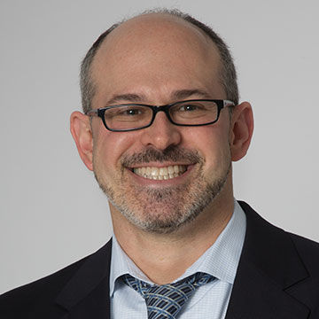 A smiling man with glasses and a gray goatee, wearing a blue shirt and tie and dark suit jacket
