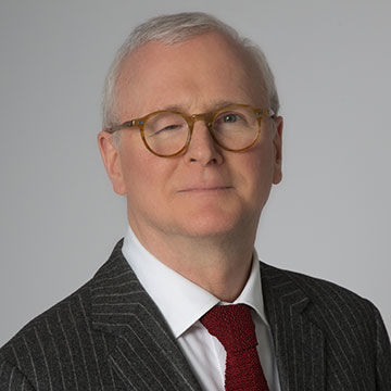 A man with short gray hair and glasses, wearing a dark gray pinstripe suit and dark red tie