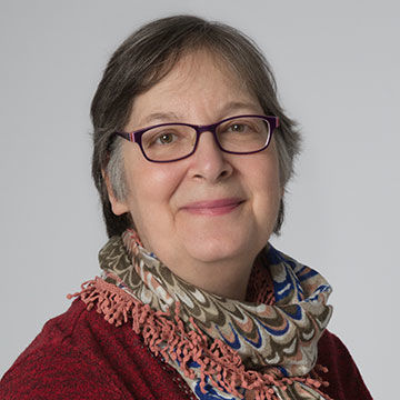 A woman with medium length graying hair and glasses, wearing a dark red sweater and multicolored crochet scarf