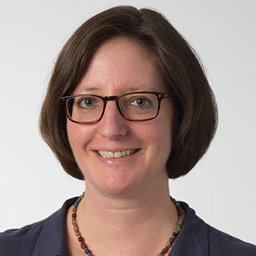 A smiling woman with medium length brown hair and glasses, wearing a purple blouse and bead necklace