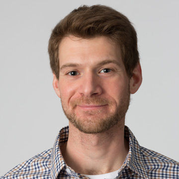 A smiling man with short brown hair wearing a plaid collared shirt.