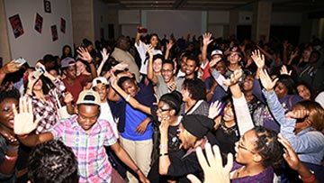 A crowd of teenagers have their hands raised in the middle of a dark dance floor