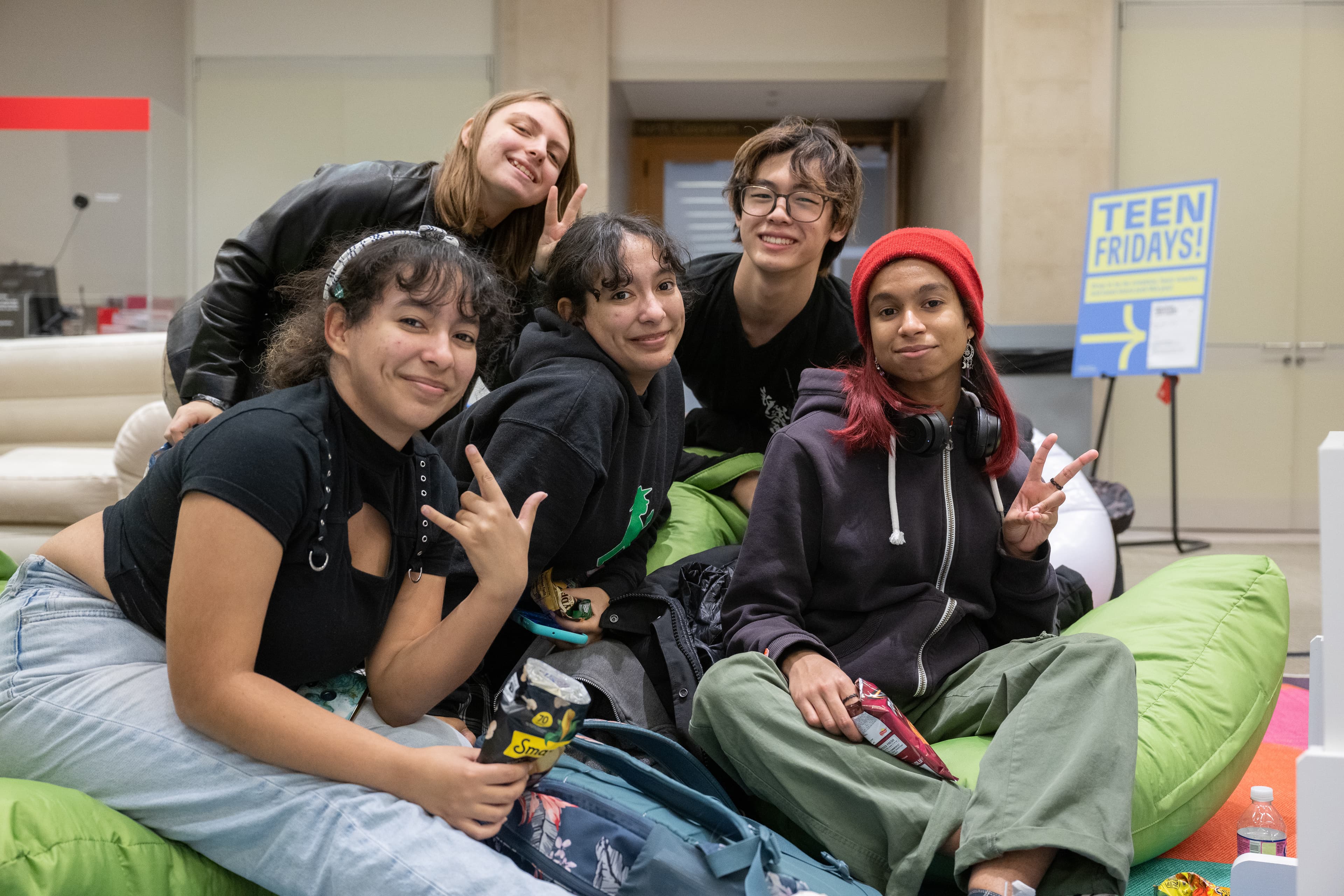 Five teens sit together on bean bag chairs. They're looking at the camera while smiling and posing. A few of the young people are making peace signs.