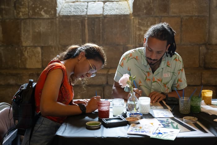 Two people paint while sitting at a table inside The Met Cloisters. The person on the left has glasses, an orange tank top, and dark hair in a ponytail. The person on the right is wearing a light blue shirt with orange flowers. On the table there is a flower in a vase, jars with water and paintbrushes, and floral paintings.     Image: Photo by Filip Wolak