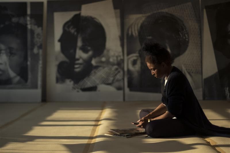A portrait of Lorna Simpson sitting on the floor in a large room. She’s sorting through magazine pages and bathed in light and shadow from a window off-frame. Large black and white prints hang behind her.